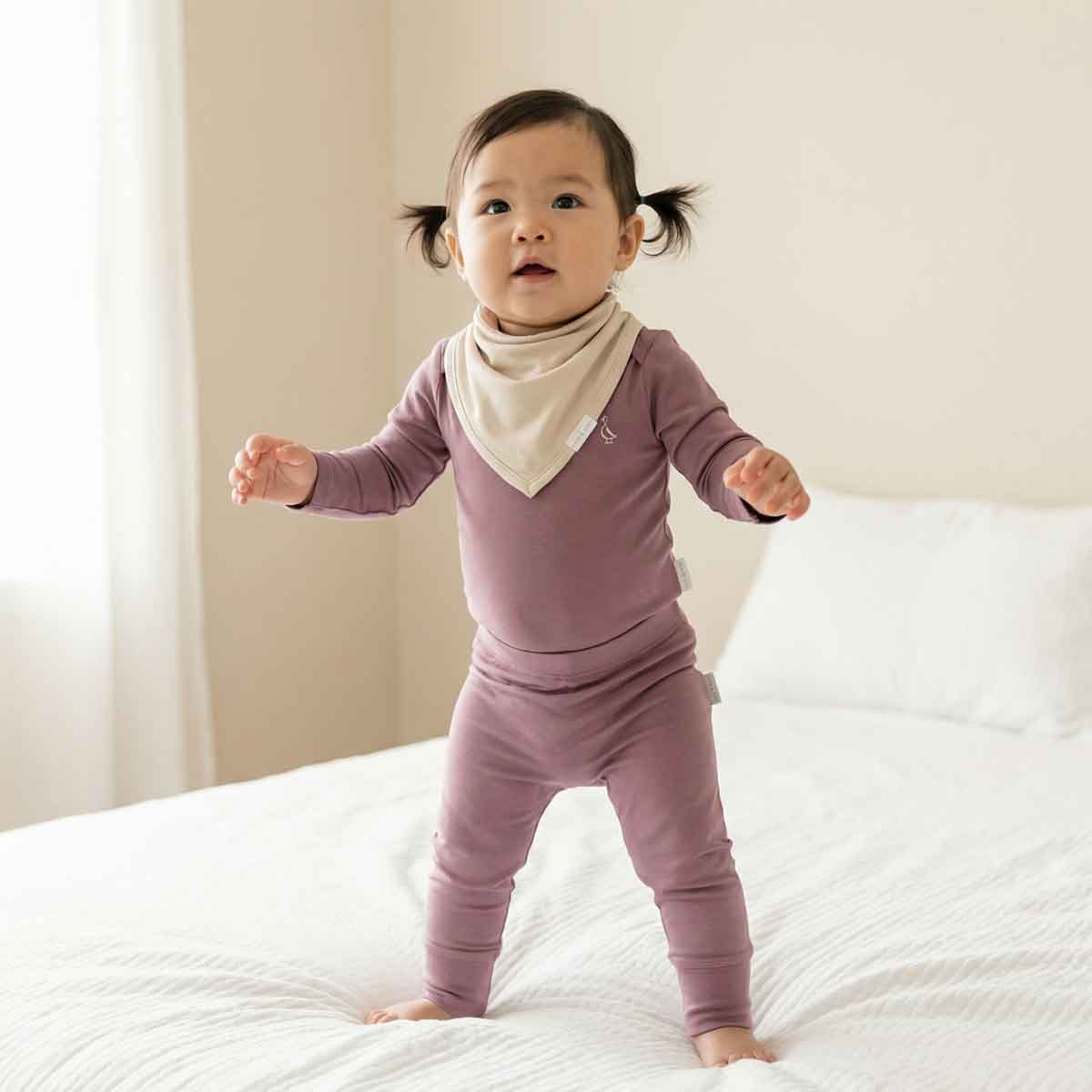 Baby in a purple outfit standing on a bed with a neutral background